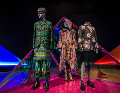 three mannequins, one dressed in green suit, one in floral hooded garment, one in black suit with pink flowers and black short pants