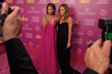 Jennifer Aniston's and Nicole Beharie's characters on The Morning Show pose in front of a pink step-and-repeat for photographers; FIT is among the logos on the background