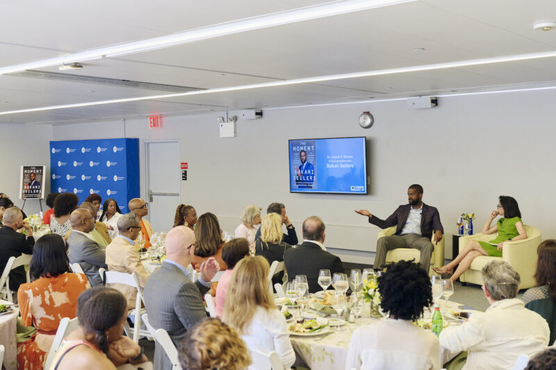 Bakari Sellers sits with President Brown in front of a large group of people at luncheon tables