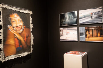 Photographs on a dark wall, on of a Black woman with a girl's mouth, four more of people in cities
