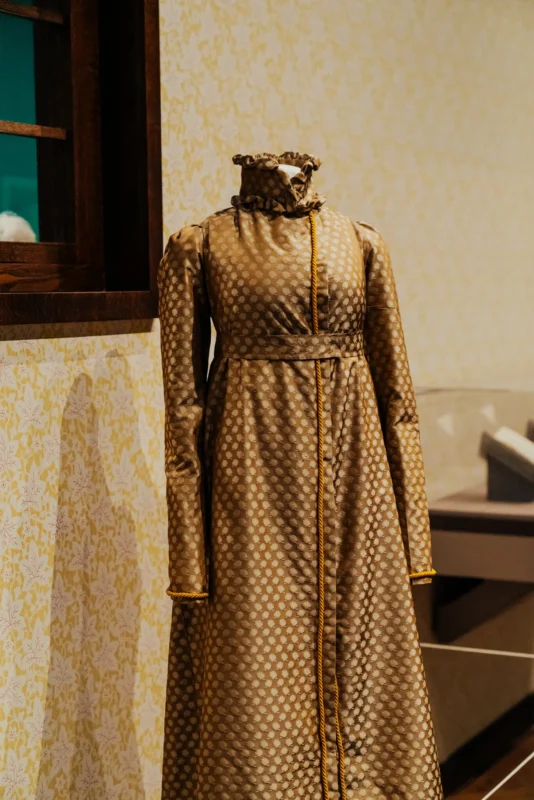 Museum mannequin displaying a brown silk coat with high collar, empire waist, and long sleeves.